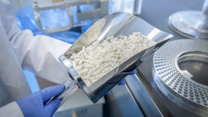 Gloved technician holding a scoop of white capsules in a pharmaceutical manufacturing facility.